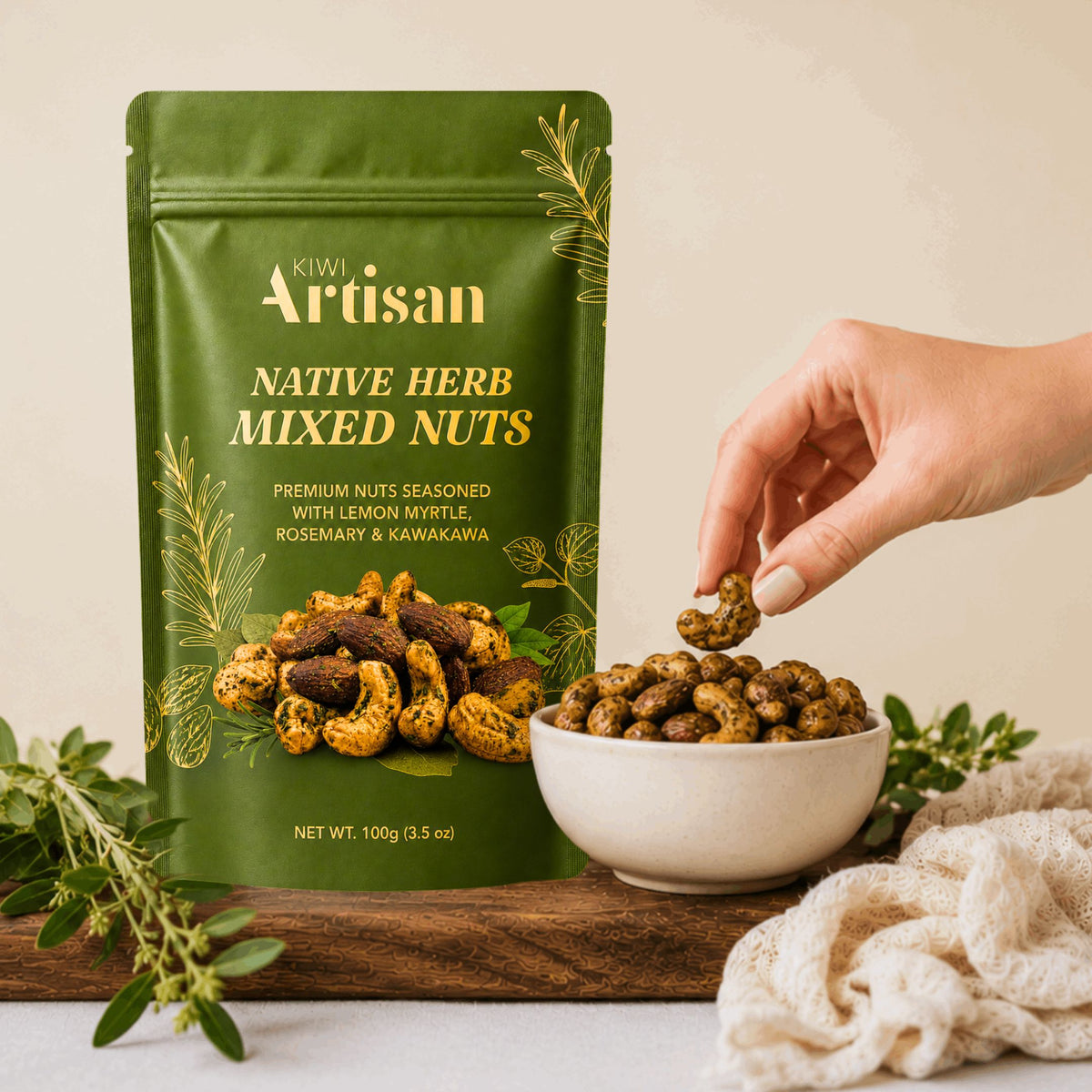 A green stand-up pouch of Kiwi Artisan Native Herb Mixed Nuts stands on a wooden board next to a white bowl filled with the nuts, with a hand reaching in to pick one up. The scene is styled with fresh herbs and a cream cloth on a light neutral background.