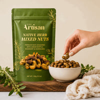 A green stand-up pouch of Kiwi Artisan Native Herb Mixed Nuts stands on a wooden board next to a white bowl filled with the nuts, with a hand reaching in to pick one up. The scene is styled with fresh herbs and a cream cloth on a light neutral background.