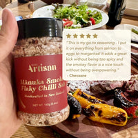 Hand holding a jar of Kiwi Artisan Manuka Smoked Flaky Chilli Salt with a background of food.