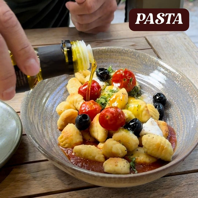Pasta dish with cherry tomatoes and olives being drizzled with black truffle  olive oil on a wooden table.